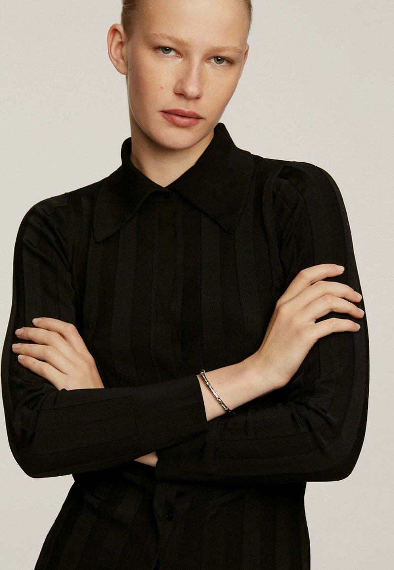 Camisa negra de manga larga con cuello y rayas verticales. Textura suave con cierre de botones. El modelo tiene los brazos cruzados, lleva una pulsera de plata.