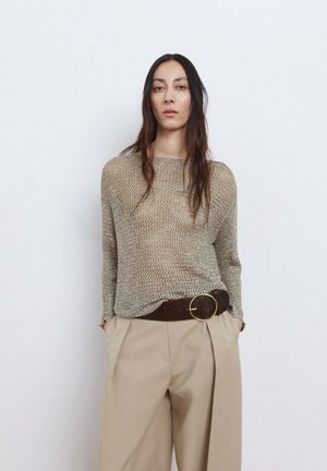 Donna con capelli lunghi e scuri che indossa un maglione beige lavorato a maglia leggero, pantaloni beige a vita alta e una larga cintura marrone con grande fibbia rotonda, con le mani nelle tasche.