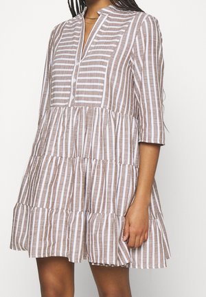 Kobieta w luźnej, warstwowej sukience w pionowe brązowo-białe paski, z rękawami do łokcia i małym dekoltem w kształcie litery V.