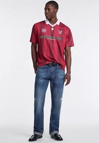 Sarkani svītraina polo krekls ar baltu apkakli, uz kura ir logotips un zīmola nosaukums. Kombinēts ar zilām nodiluma efektu džinsām un melniem apaviem.