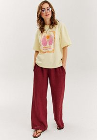 Svetle žluté oversized tričko s grafickým potiskem, na kterém jsou ruce a slunce, v kombinaci s volnými burgundskými kalhotami a černými žabkami.