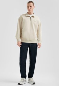 Felpa zip-up beige con polsini e orlo a costine, abbinata a jeans denim scuri e sneakers bianche con dettagli grigi.