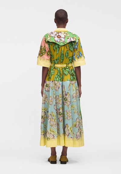 Personne portant une robe midi colorée à imprimé floral avec manches courtes, une ceinture et des poignets jaunes, debout de dos sur un fond uni.