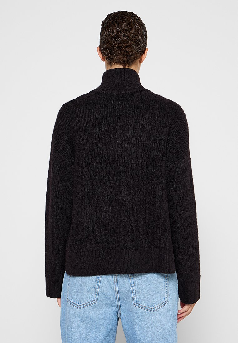 Suéter negro de punto con un cuello alto, hombros caídos y un corte relajado. Textura suave y diseño acanalado, combinado con jeans de algodón azul claro.