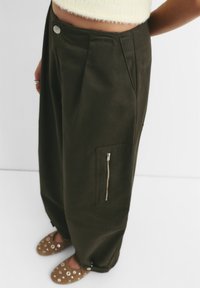 Donkergroene wijde broek met voorplooien, zijzak en zilveren knoopsluiting. Gladde stof met een relaxte pasvorm.