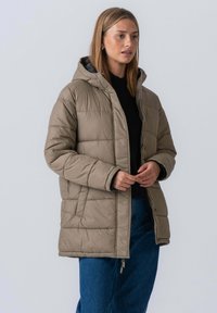 Khaki pufferjakke med hætte, kviltet design, forlommer og lynlåsclosing, båret over en sort sweater og blå jeans.
