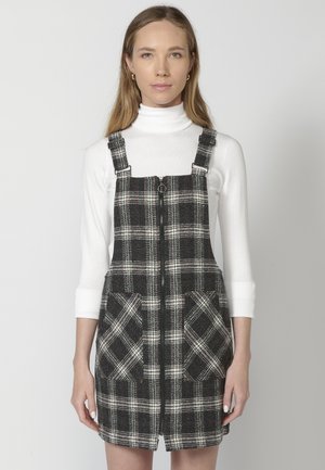Robe salopette à carreaux noir et blanc avec fermeture éclair devant, dotée de deux grandes poches avant et de bretelles ajustables, portée sur un col roulé blanc.