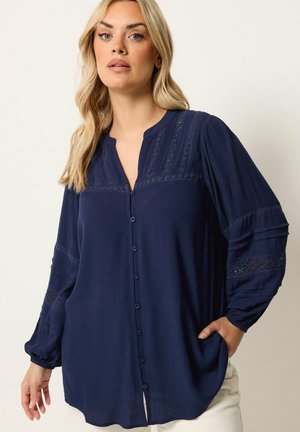 Kvinde iført en mørkeblå, langærmet skjorte med knapper og blonde detaljer på ærmer og skuldre, kombineret med lyse bukser.