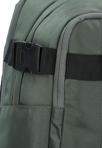 Mochila de tela verde con una superficie texturizada, hebilla negra y bolsillos laterales. Estructura resistente con costuras reforzadas y correas ajustables.