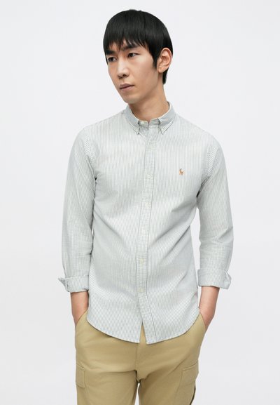 Světle zelená a bílá pruhovaná košile na knoflíky s límcem, na hrudi s malým vyšívaným logem; látka vypadá hladká a lehká.