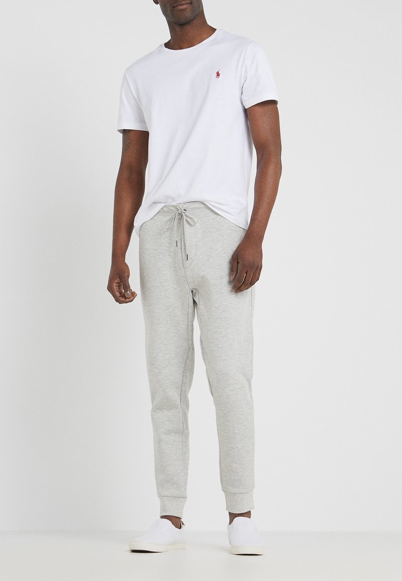 Valkoinen lyhythihainen T-paita, jossa punainen merkki, yhdistetty vaaleanharmaisiin collegehousuihin, joissa on nyöritysvyötärö ja käännetyt lahkeet. Valkoiset slip-on-kengät.