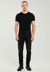 Tânăr în picioare, purtând un tricou negru, blugi negri și cizme negre, cu fața spre înainte, pe un fundal simplu și deschis la culoare.