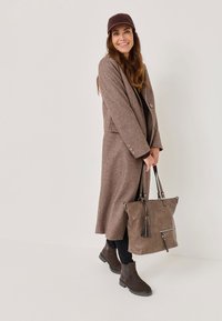 Brązowy wełniany płaszcz, ciemna bluzka, czarne dżinsy, brązowe botki za kostkę oraz szara torba z frędzlami, wszystko na neutralnym tle.