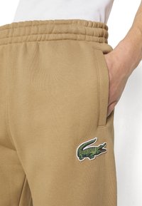 Beige Jogginghosen aus strukturiertem Material, mit einem gestickten grünen Krokodil-Logo an der rechten Tasche und einem elastischen Bund.