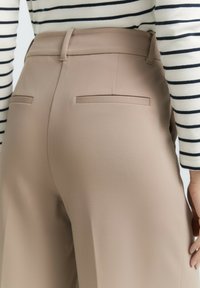 Beige tailored trousers with a smooth texture, featuring two back pockets and a straight-leg design, paired with a striped top.
