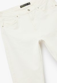 Białe jeansy o tapered fit z pięcioma kieszeniami, wykonane z denimu, z widocznym przeszyciem i srebrnymi elementami. Metka wskazuje rozmiar W32 L32.