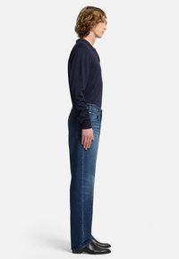 Granatowa, długorękawowa koszulka polo, niebieskie dżinsy, slim fit, prosta nogawka, delikatne przetarcia, noszone z czarnymi skórzanymi butami. Widok z boku.