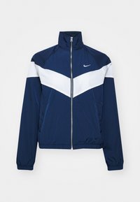 Námořnický modrý větrovka s bílým chevronovým designem, vysokým límcem, zipovým zapínáním a elastickými manžetami. Vyrobeno z lehké, hladké tkaniny.