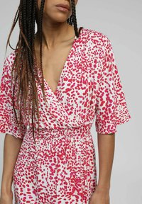 Robe blanche avec un motif de taches roses, design en V croisé, manches courtes et taille élastique. Tissu texturé avec une coupe ample.