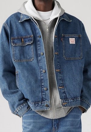 Homme portant une veste en jean superposée sur un sweat à capuche gris zippé et un t-shirt blanc, mains dans les poches, tenue décontractée.