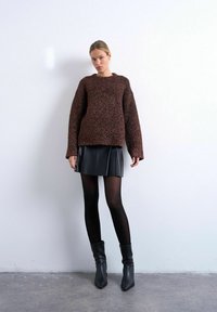 Femme portant un pull marron texturé, une mini-jupe en cuir noir, des collants noirs et des bottines noires, debout contre un mur blanc uni.