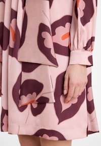 Händchen ruht neben einem pinken Kleid mit großen lila und orangefarbenen Blütenmotiven, wobei der hochgekrempelte lange Ärmel und die Stoffstruktur im natürlichen Licht sichtbar sind.