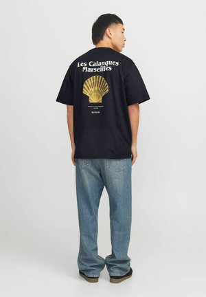 Man draagt een zwart T-shirt met een gele schelpafbeelding en de tekst "Les Calanques Marseilles" op de rug, gecombineerd met losse lichtblauwe spijkerbroek.