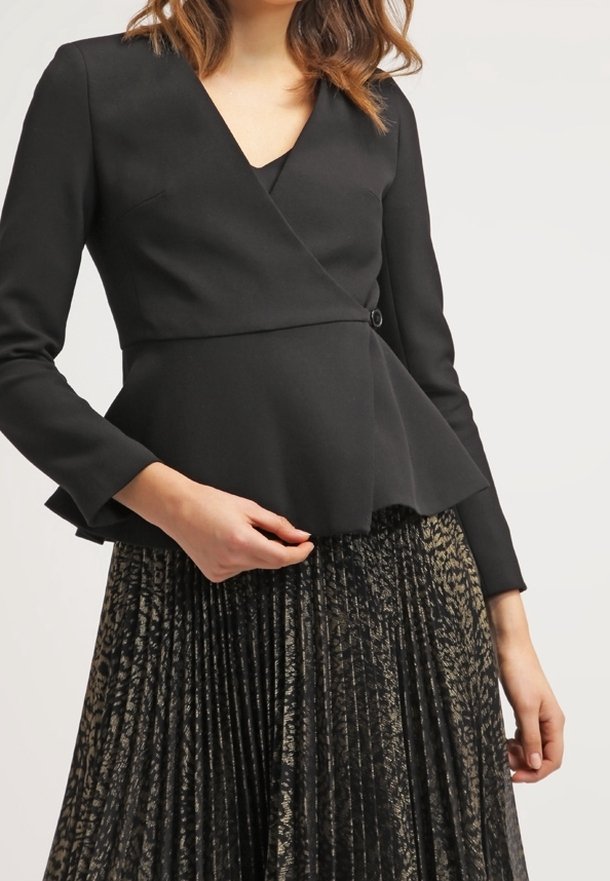 Femme portant un blazer noir cintré style portefeuille avec un ourlet à basque et une jupe plissée foncée avec un imprimé animal discret.