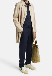 Trench-coat beige porté par-dessus un polo bleu marine, un pantalon foncé et des baskets blanches avec des touches de vert. Un sac fourre-tout marron complète la tenue.