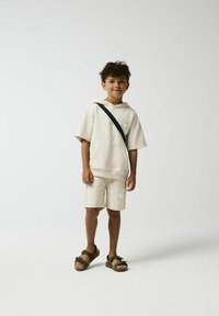Beige hoodie met korte mouwen, bijpassende shorts en donkere sandalen; beschikt over een grafisch patroon aan de voorkant en een schouderband.
