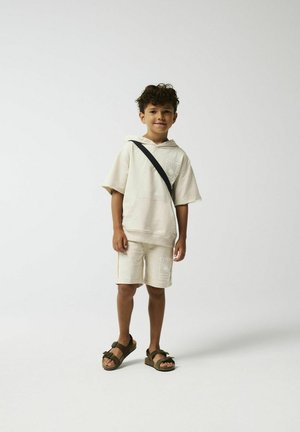 Felpa beige con cappuccio e maniche corte, pantaloni corti abbinati e sandali scuri; presenta un motivo grafico frontale e tracolla.