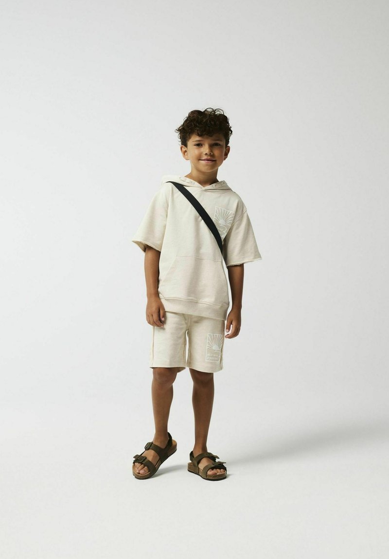 Beige hoodie met korte mouwen, bijpassende shorts en donkere sandalen; beschikt over een grafisch patroon aan de voorkant en een schouderband.