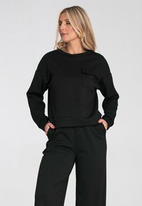 Czarny sweter wykonany z miękkiego materiału, z okrągłym dekoltem, długimi rękawami i przednią kieszenią. W połączeniu z pasującymi czarnymi spodniami.