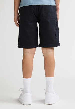 Pantalones cortos cargo azul marino oscuro con dos bolsillos traseros, dobladillos terminados y costuras reforzadas. Combinados con zapatillas blancas y calcetines de canalé.