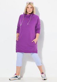 Langer lila Hoodie mit Kapuze und Kordelzügen. Verfügt über seitliche Taschen, gerippte Bündchen und wird mit hellblauen, cropped Leggings und deportivas Schuhen kombiniert.