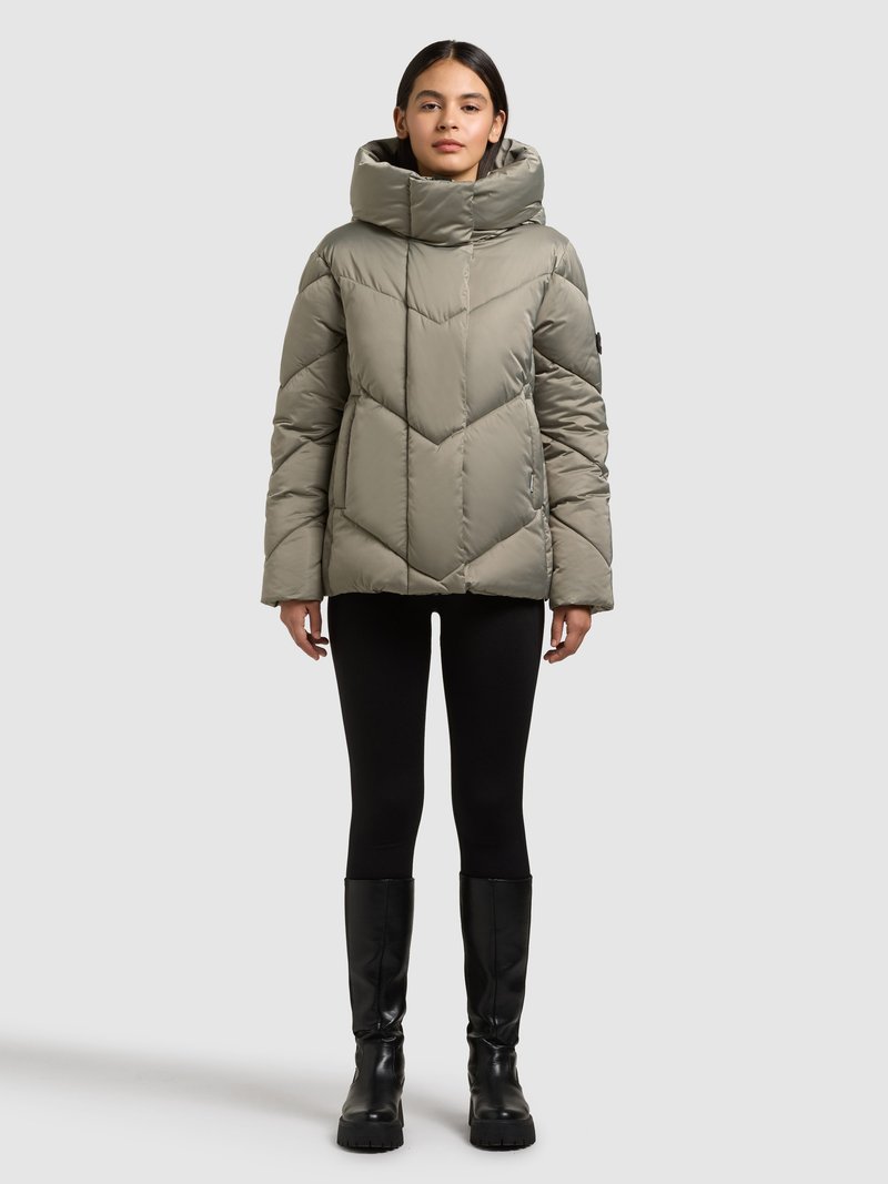 Polsterēta kapučveida jaka matētā olīvu zaļā krāsā ar šķērsenisku rakstu, ar augstu apkakli, rāvējslēdzēja aizdari un sānu kabatām, kopā ar melnām legingiem un zābakiem.