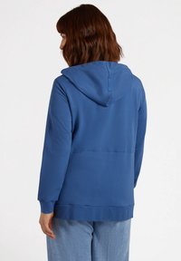 Blue hooded sweatshirt with a smooth texture, kangaroo pocket, and ribbed cuffs, paired with light blue loose-fitting jeans.
