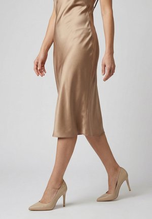 Kvinde går iført en skinnende beige midi-kjole og matchende beige højhælede pumps mod en ensfarvet baggrund.