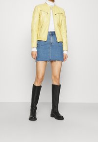 Giacca di pelle gialla con tasche con zip, gonna di jeans aderente con orlo sfrangiato e stivali neri in pelle al ginocchio.