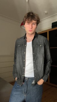 Camicia button-up in denim nera con due tasche sul petto, abbinata a una t-shirt bianca e jeans blu. Ambiente interno, illuminazione soffusa.