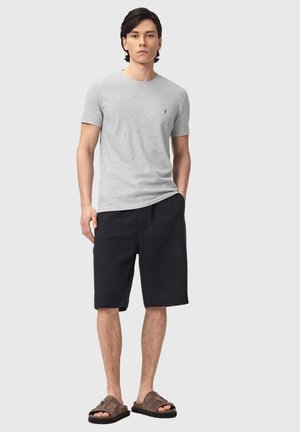 Junger Mann steht und trägt ein graues T-Shirt, schwarze kniehohe Shorts und braune Sandalen vor hellem grauem Hintergrund.