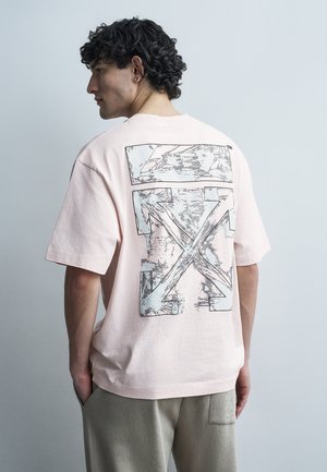 Mann mit lockigem Haar trägt ein hellrosa übergroßes T-Shirt mit großem abstraktem Grafikdesign auf dem Rücken, steht vor einem schlichten grauen Hintergrund.