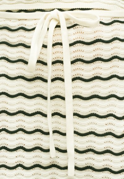 Κοντινό πλάνο υφάσματος πλεκτού σε κρεμ χρώμα με μαύρες κυματιστές οριζόντιες ρίγες και δεμένο φιόγκο από κορδόνι σε κρεμ απόχρωση στο πάνω κέντρο.