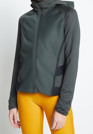 Osoba nosząca ciemnoszarą bluzę z kapturem na zamek z teksturowanymi panelami z przodu oraz jaskrawożółte legginsy, stojąca na tle jednolitego tła.