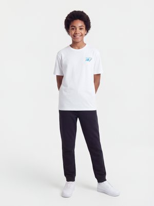 Criança com cabelo cacheado sorri vestindo uma t-shirt branca de manga curta, calças pretas e ténis brancos contra um fundo liso.