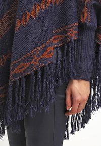 Marinblå och rostfärgad mönstrad stickad poncho med fransar, bärs över svarta byxor, med en närbild av en hand som vilar vid kroppen.