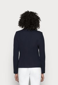 Blazer blu navy su misura con spalle strutturate, maniche lunghe e chiusura a un bottone. Presenta un tessuto testurizzato e una fessura posteriore per una maggiore libertà di movimento.