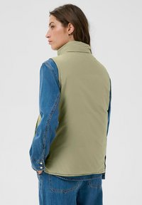 Frau trägt eine hellgrüne ärmellose Weste über einer blauen Jeansjacke und Jeans, schaut nach links mit den Händen in den Taschen, einfarbiger Hintergrund.