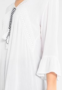 Blusa bianca caratterizzata da un tessuto testurizzato, scollatura a V, bordo in pizzo e dettaglio con laccio e nappine. Maniche a campana con dettagli arricciati.