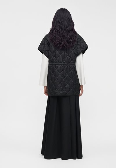 Femme aux longs cheveux foncés portant une veste matelassée noire à manches courtes par-dessus un haut blanc à manches longues et une longue jupe noire, vue de dos.
